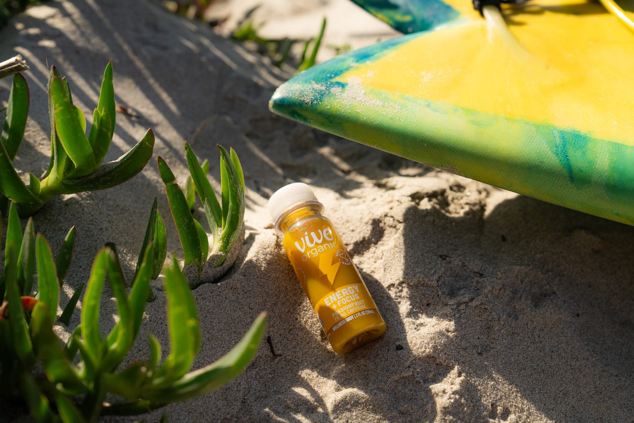 Vive Organic energy shot nestled in beach sand between ice plant and a yellow surfboard in morning sun