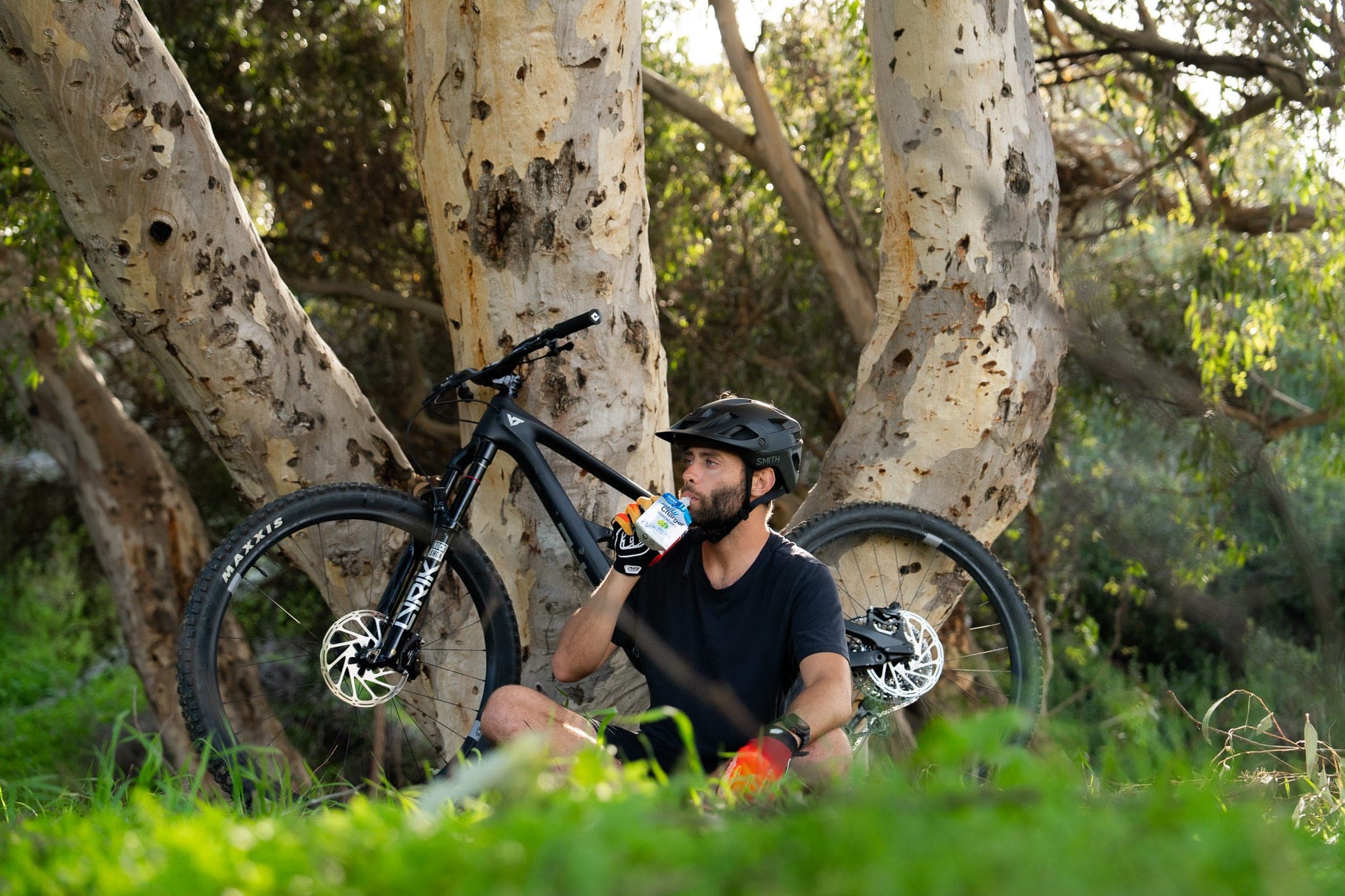 Mountain biker resting under eucalyptus trees, sipping a recovery drink beside his bike in golden light