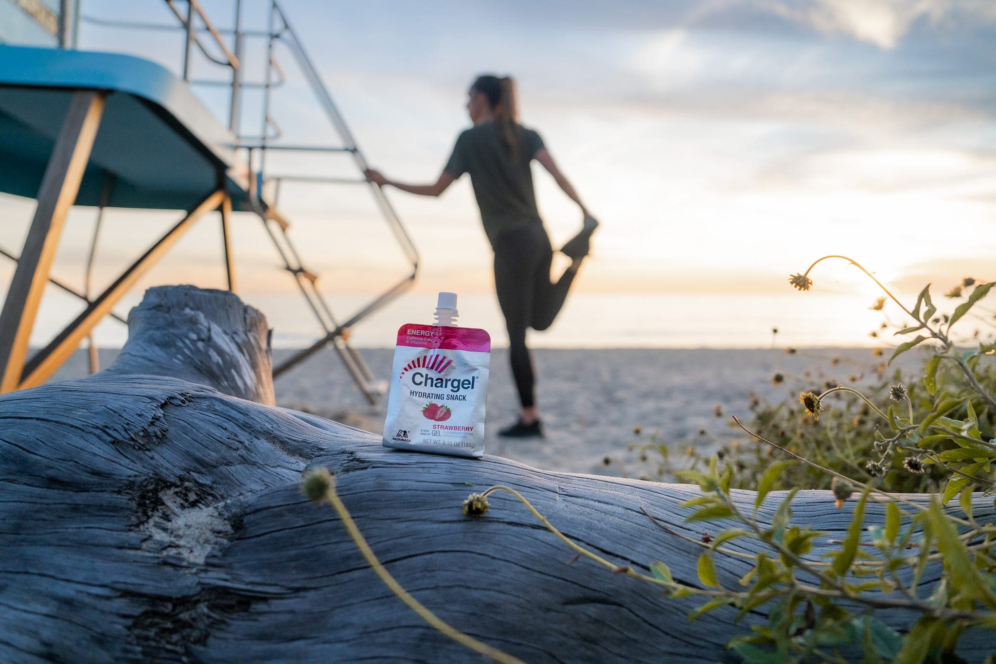 Chargel energy gel pouch propped on driftwood while a runner stretches by a lifeguard tower at sunrise