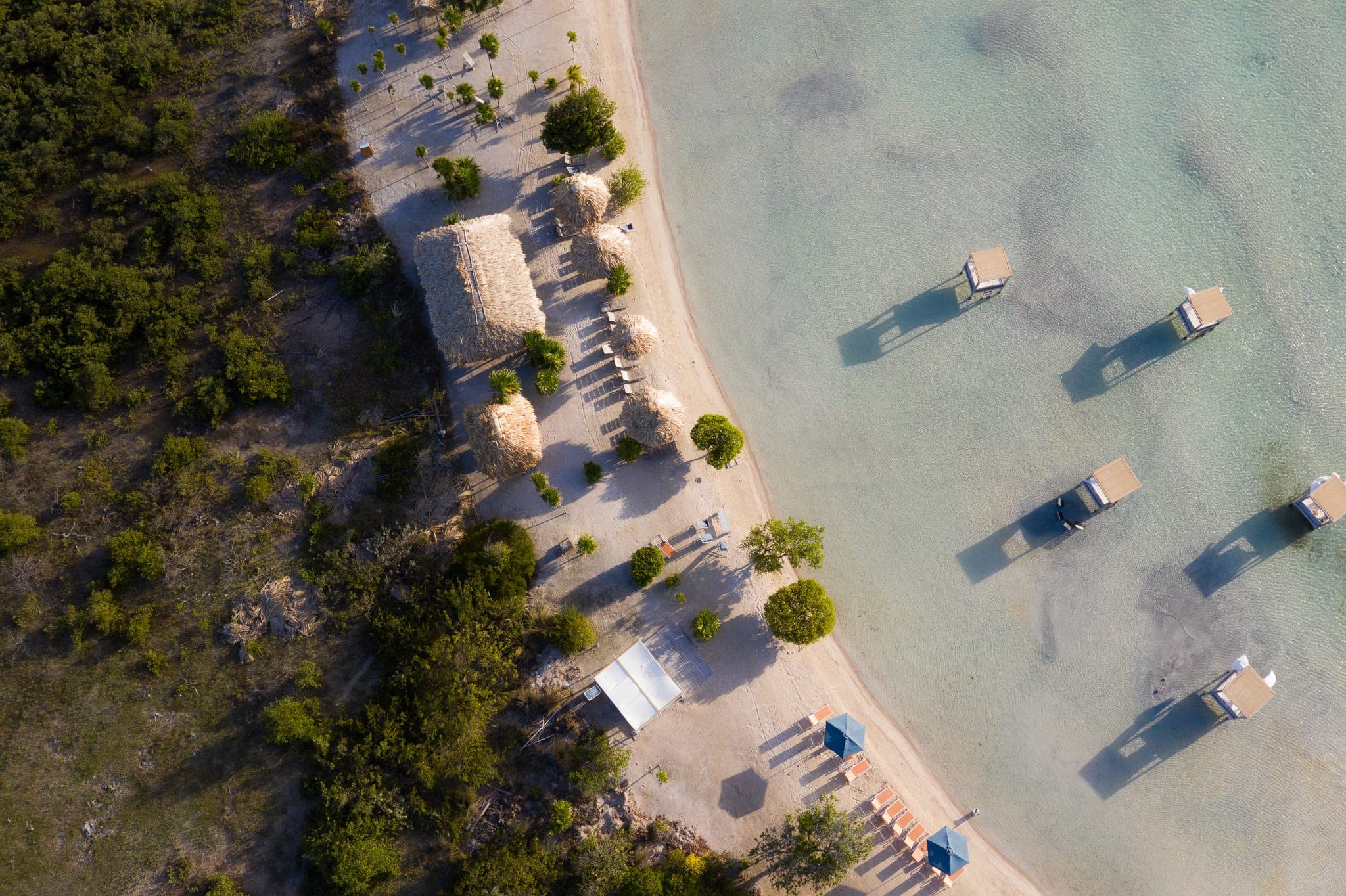Aerial view of a beach club with thatched palapas and stilted cabanas in clear shallow water