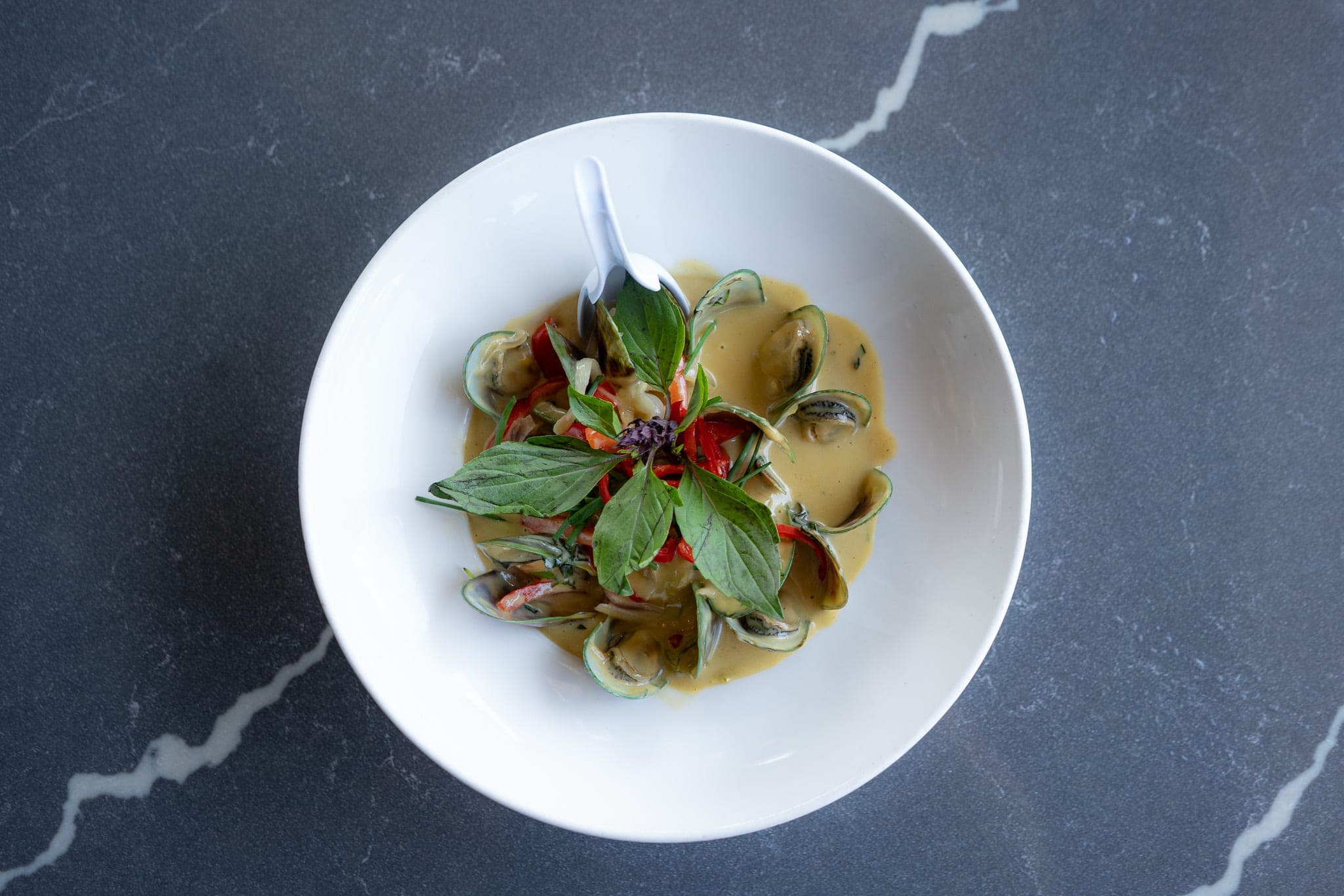 Overhead view of green-lipped mussels in coconut curry topped with Thai basil, served in a white bowl on marble