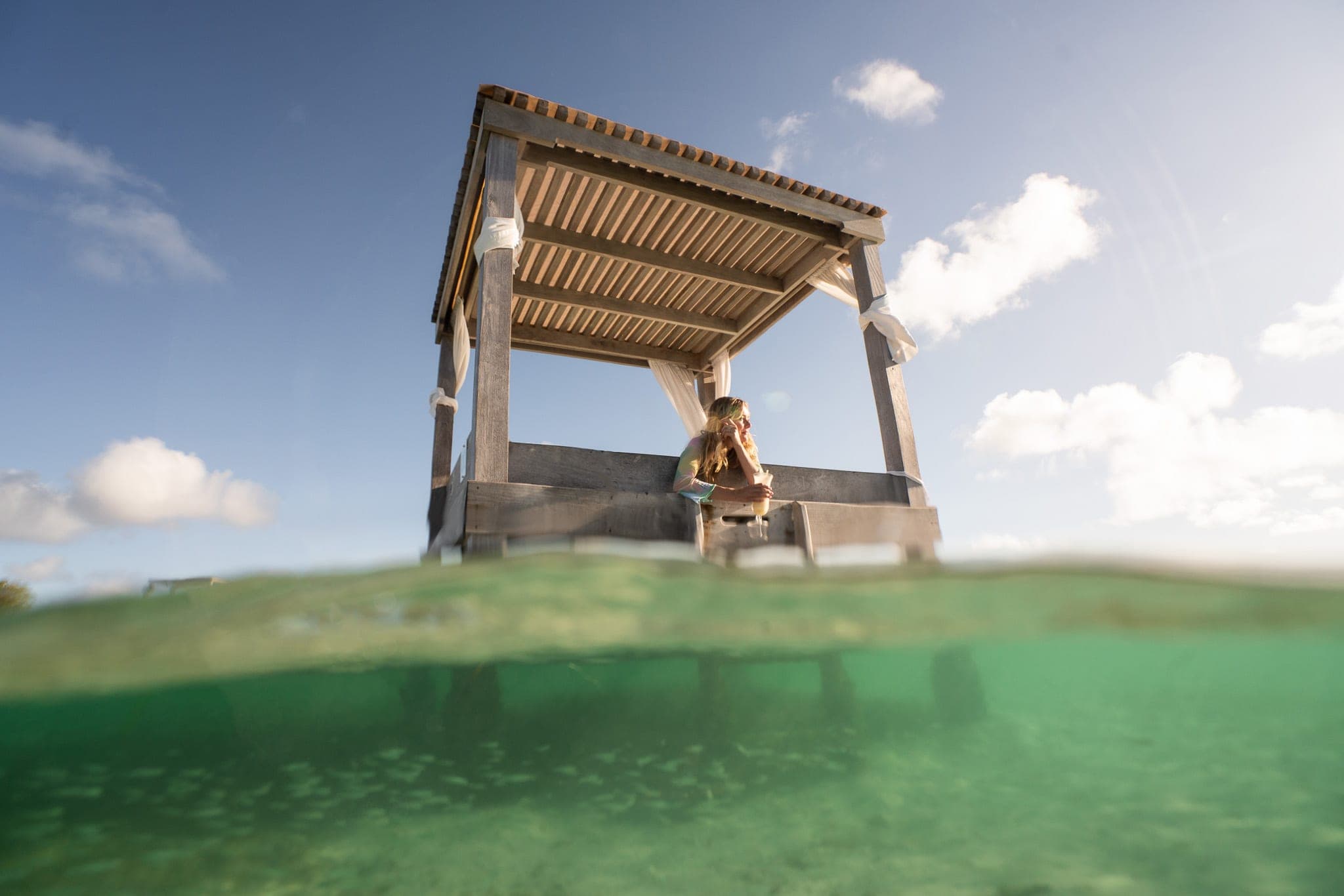 Woman enjoying a drink in a stilted overwater cabana, photographed half-underwater in a green lagoon
