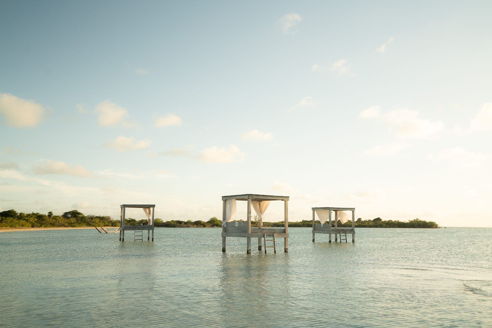 Three canopied daybeds standing on stilts in a shallow, glassy lagoon under a soft evening sky