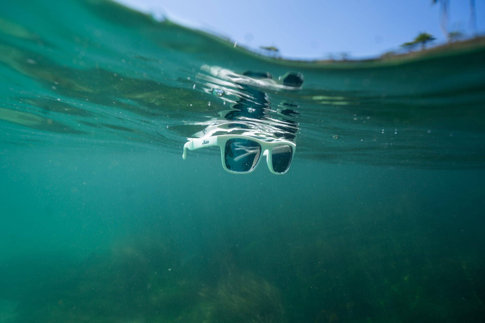 Pair of white sunglasses floating just beneath the surface of clear green ocean water