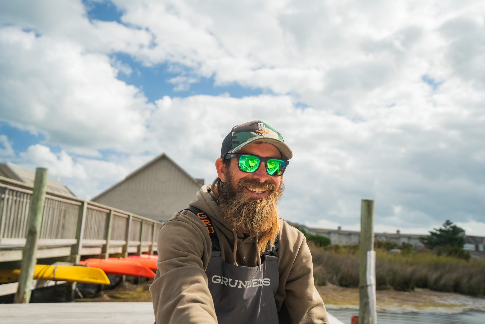 Smiling bearded fisherman in a camo cap, mirrored sunglasses, and waders on a dock lined with kayaks