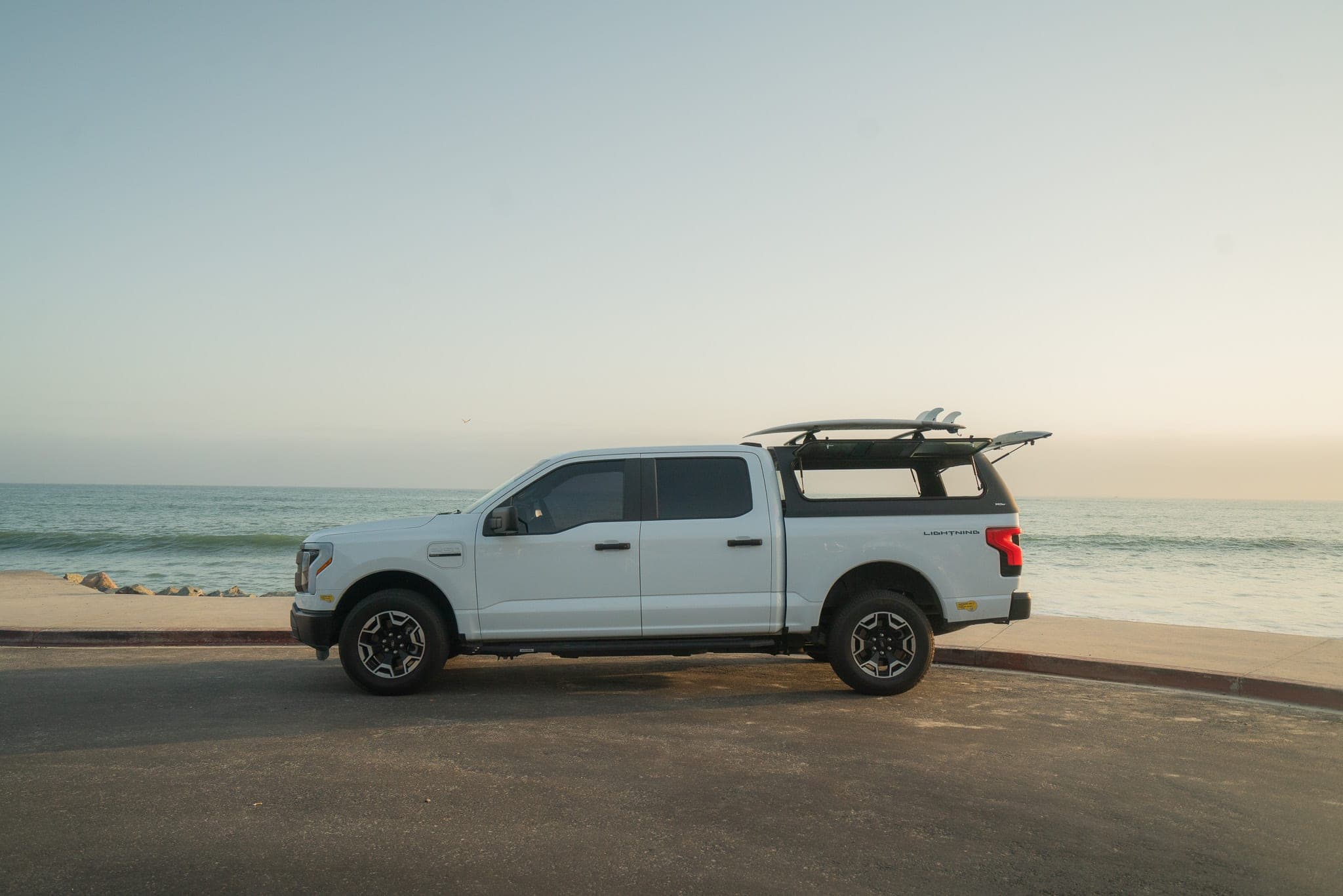 White electric pickup truck with a surfboard on its camper rack parked beside a calm ocean at dusk