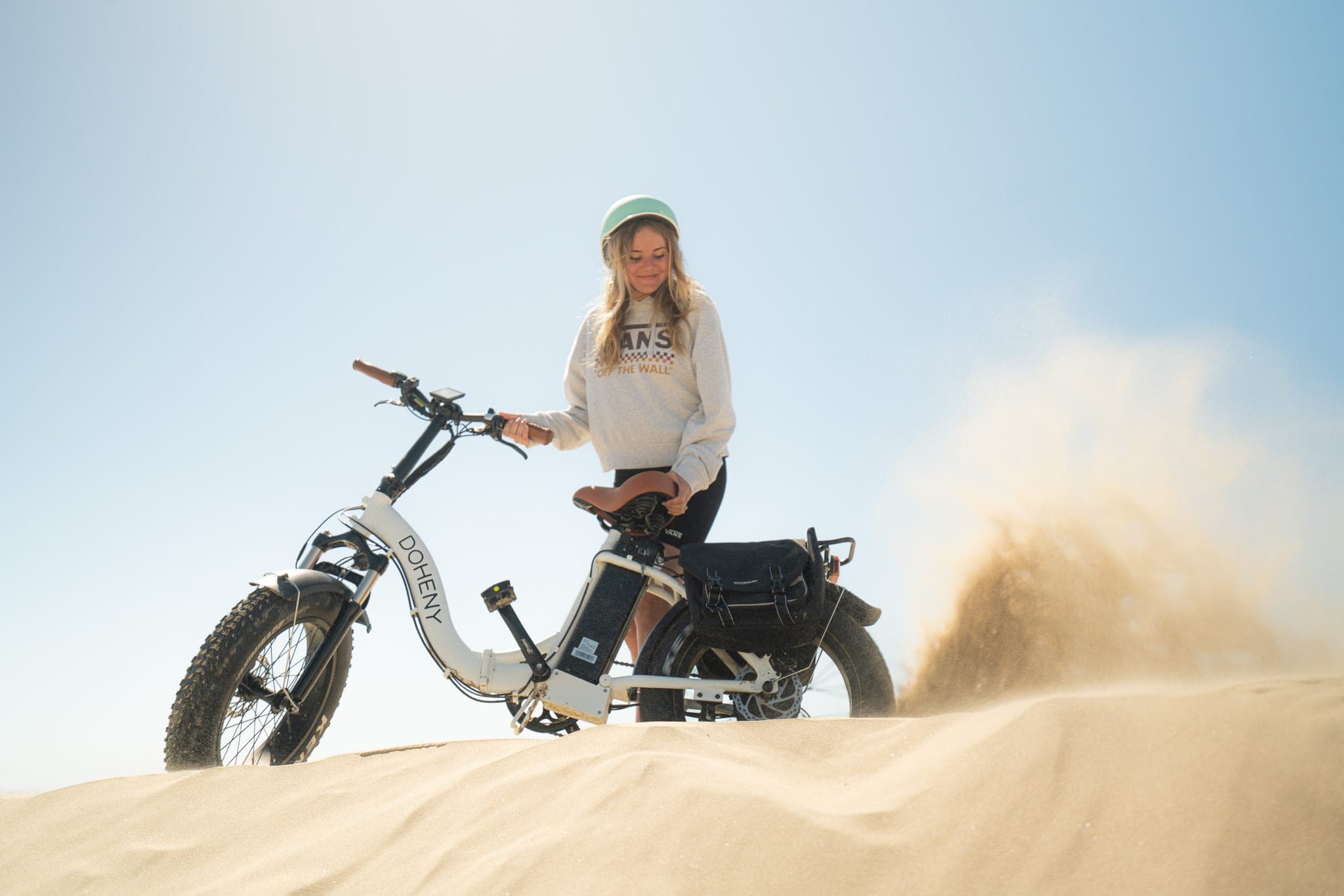 Woman in a mint helmet standing with a fat-tire e-bike on a sand dune as sand kicks up
