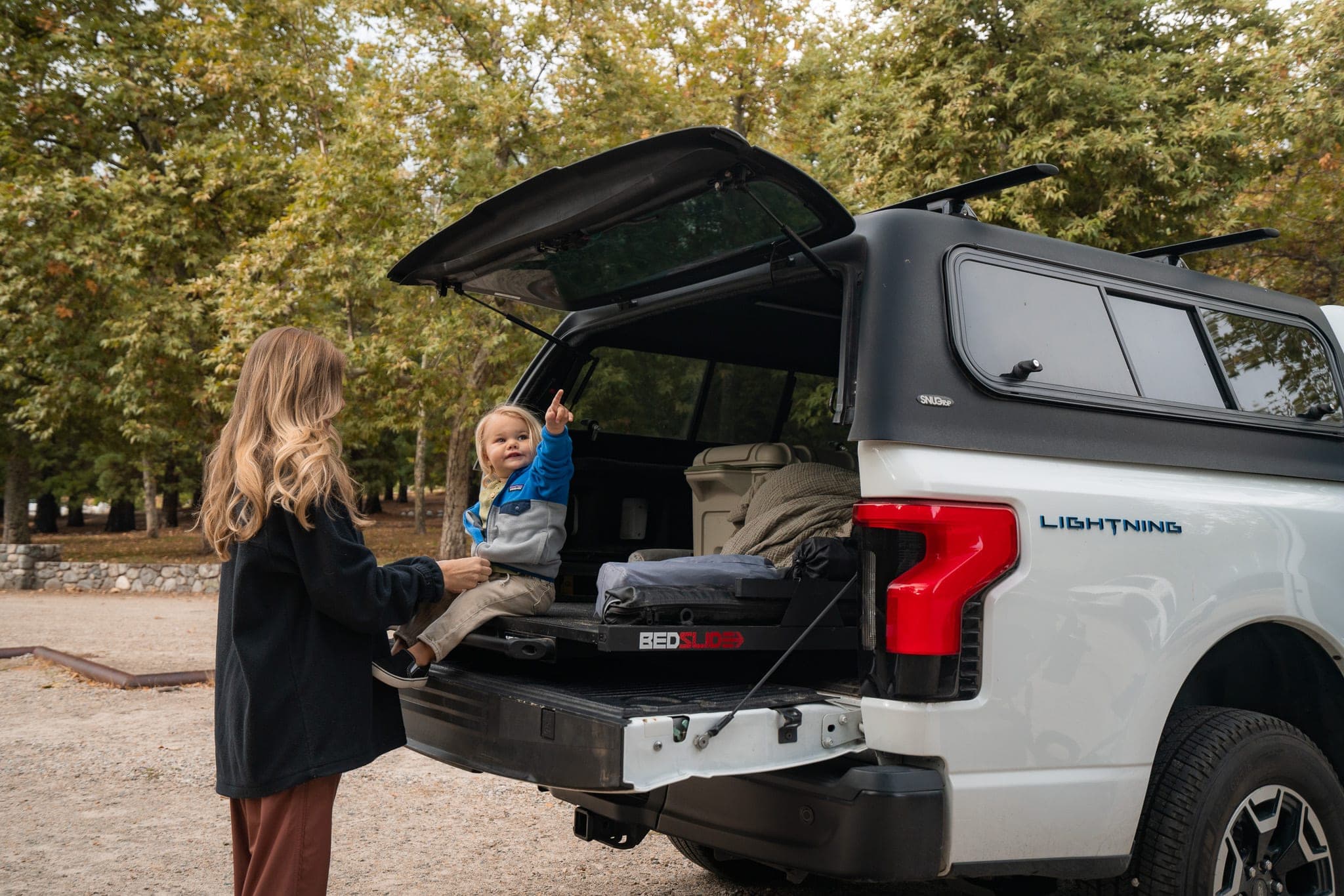Mother helping her toddler on the tailgate of a camper-topped electric pickup under autumn trees