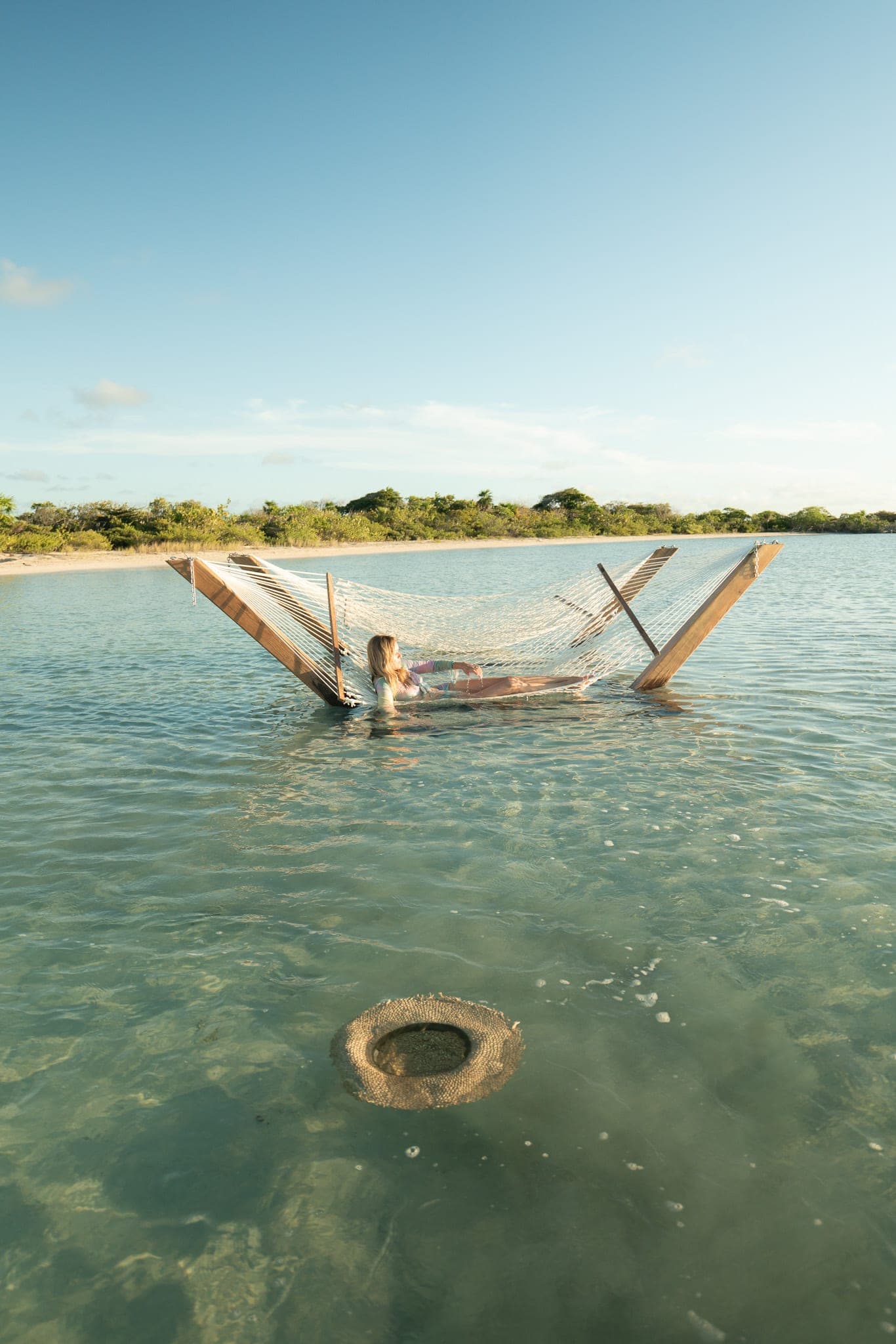 Woman relaxing in a rope hammock strung over clear shallow lagoon water, a straw hat drifting below