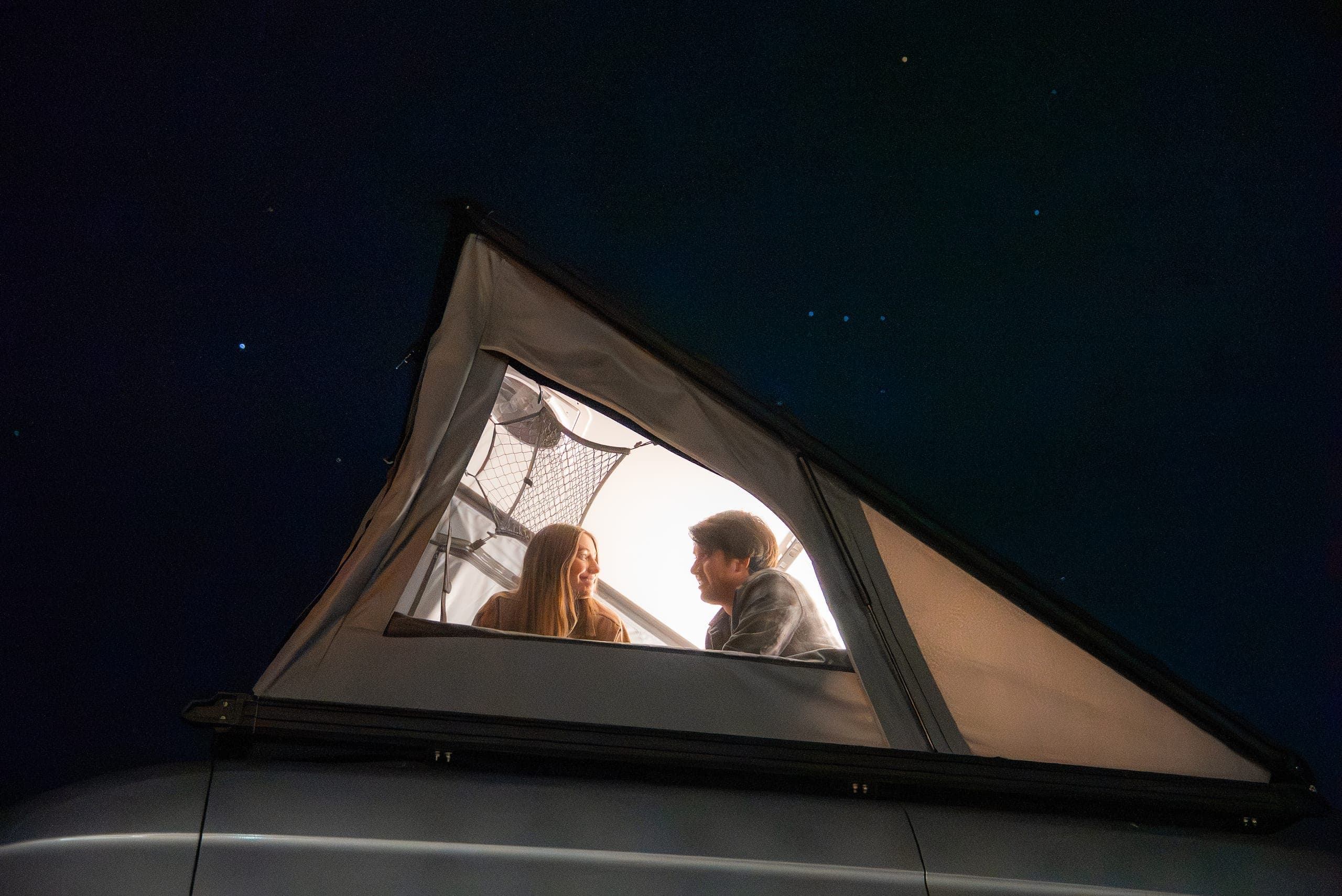 Couple smiling inside a lit rooftop tent beneath a starry night sky