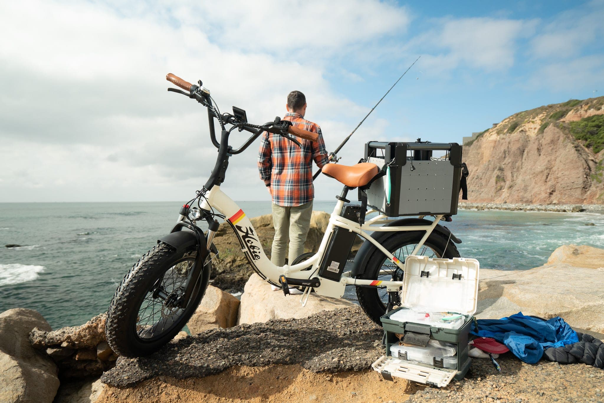 Angler casting from coastal rocks beside a fat-tire e-bike loaded with tackle boxes