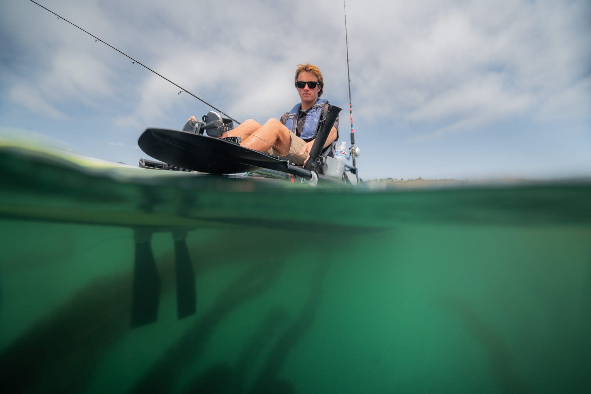 Split over-under view of a man pedaling a fishing kayak with rods against a cloudy sky
