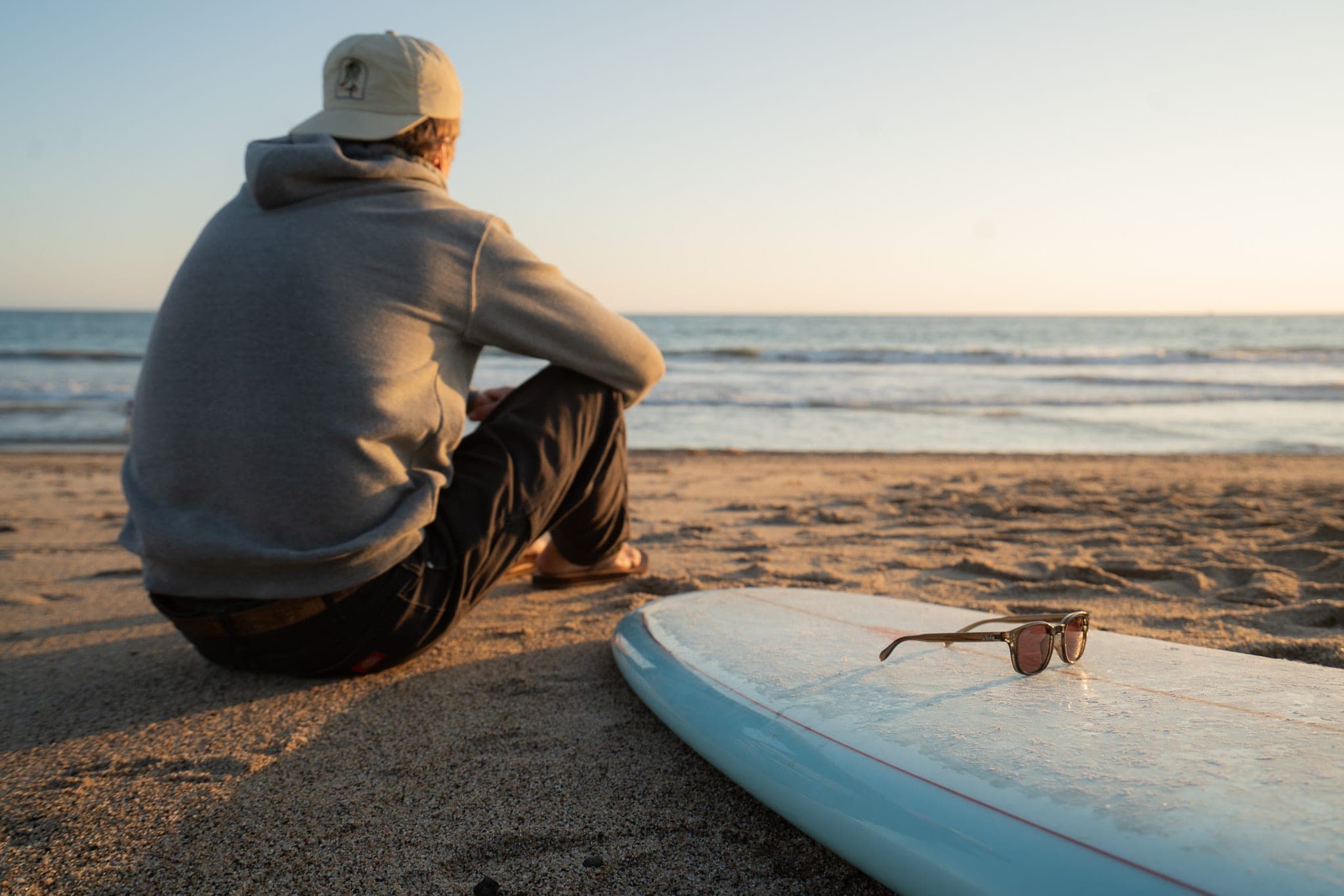 Man in a hoodie sitting on the sand at golden hour beside a longboard with sunglasses resting on it