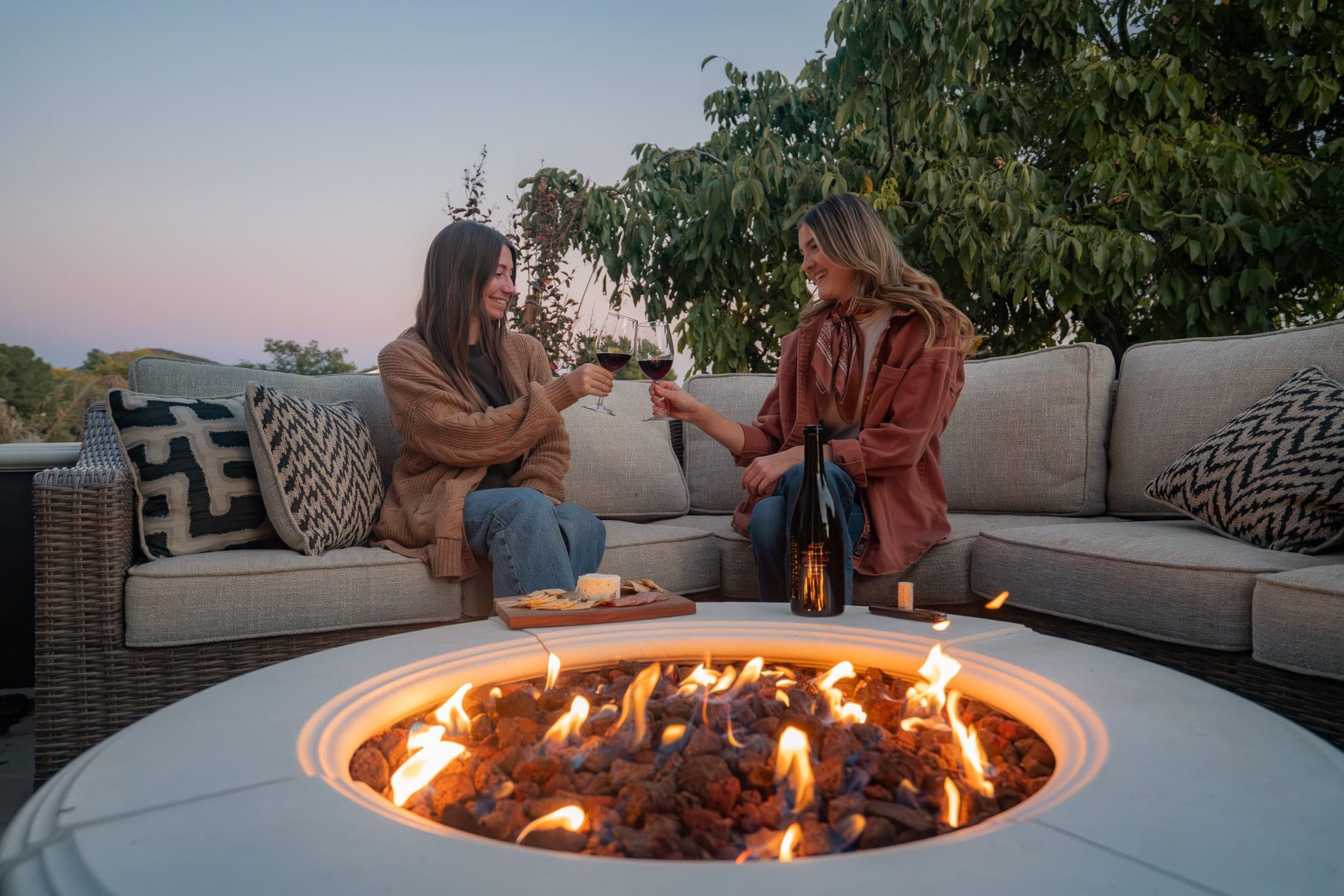 Two friends toasting glasses of red wine around a glowing fire pit at dusk