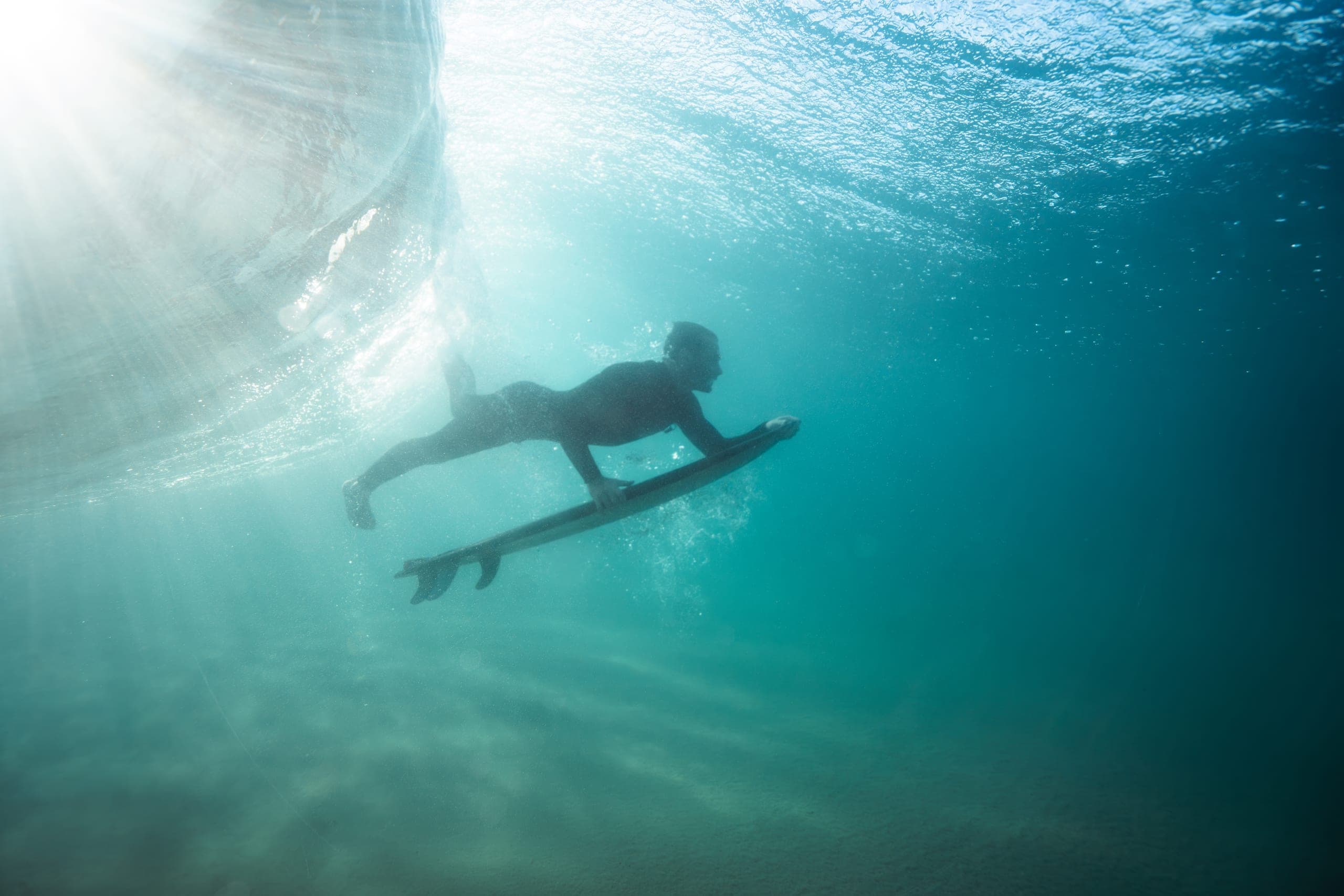 Underwater view of a surfer duck-diving his board beneath a breaking wave in sunlit turquoise water