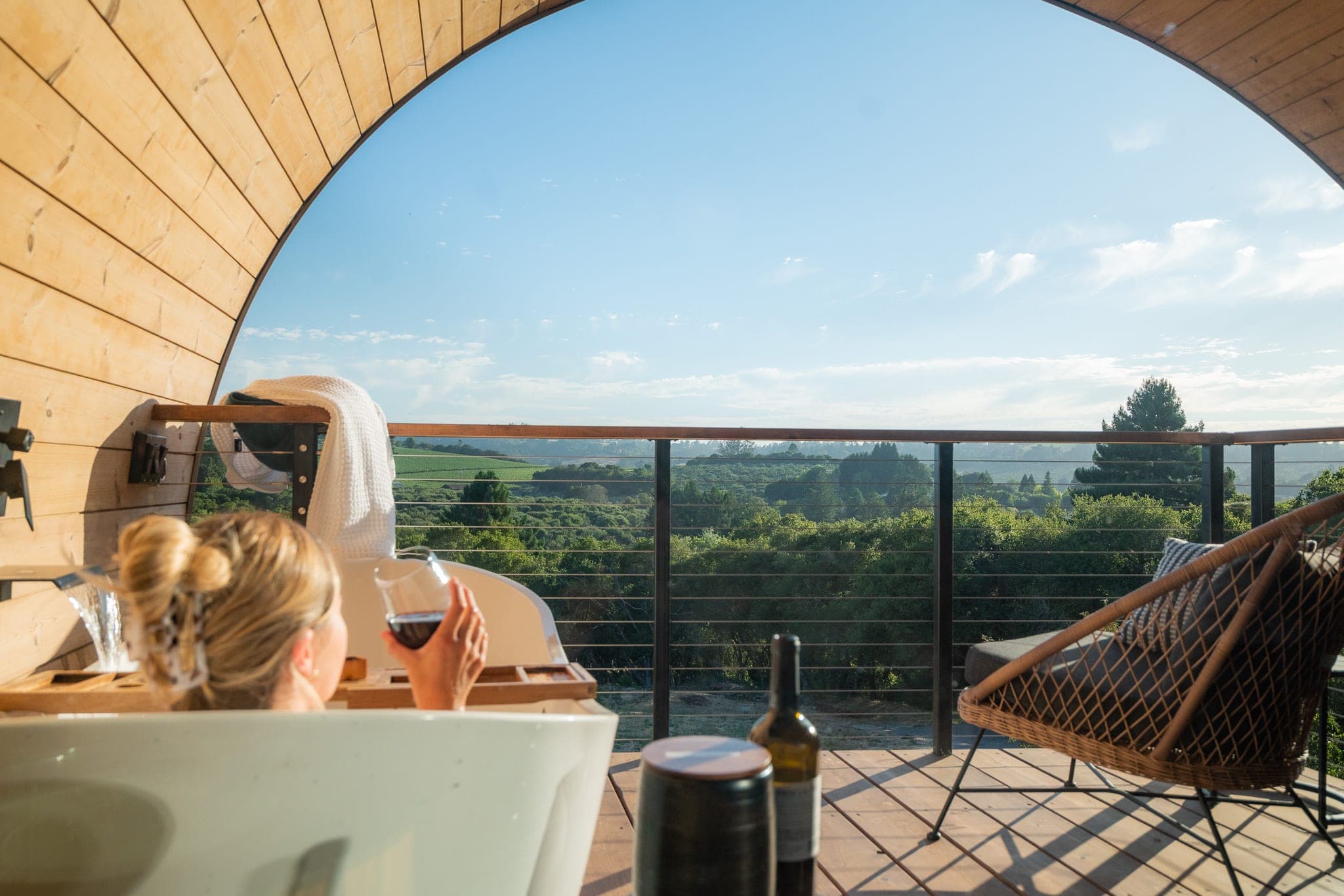 Woman sipping wine from a soaking tub on a barrel-cabin balcony overlooking vineyard hills
