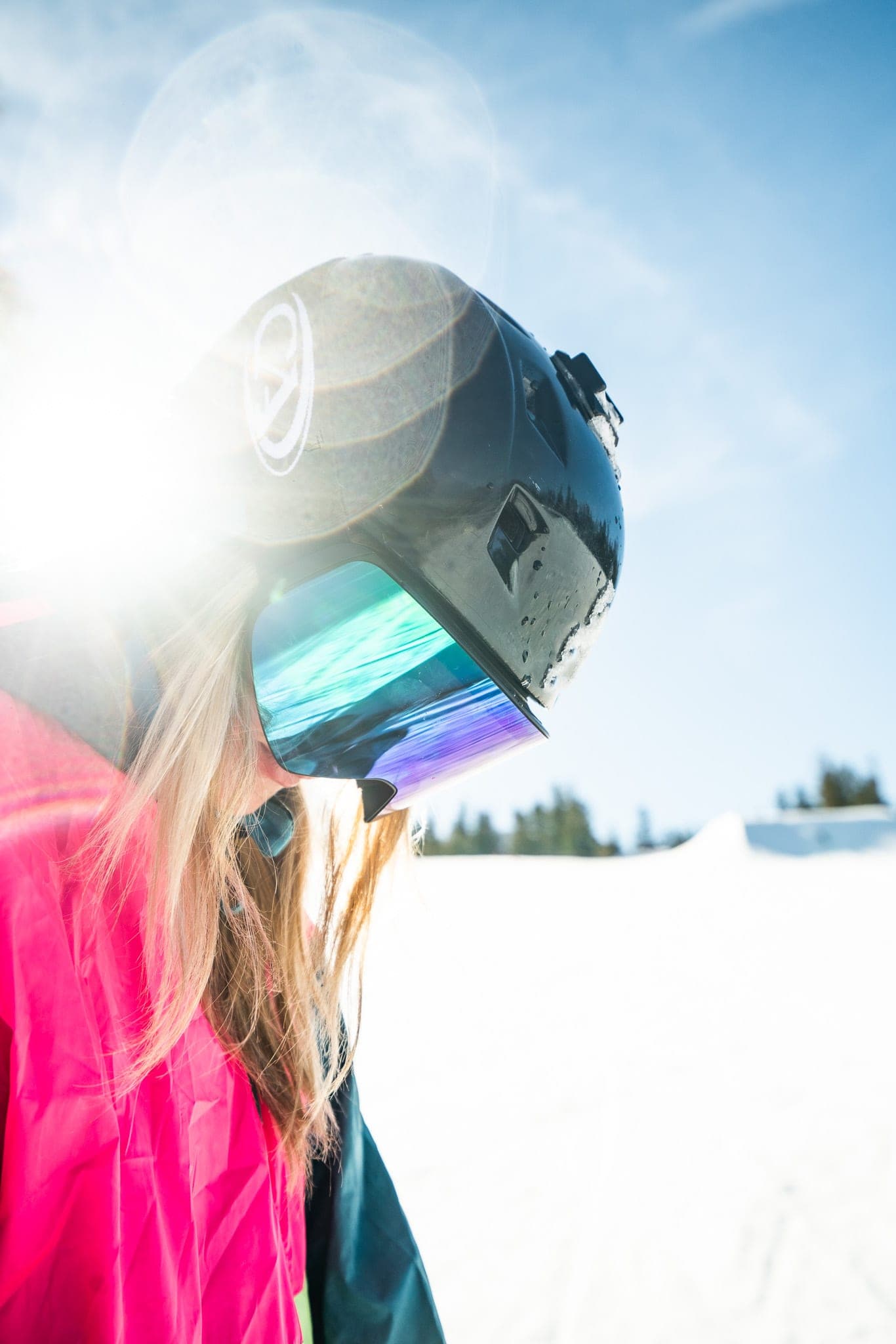 Snowboarder in a black helmet and iridescent goggles looking down in bright sun flare on a snowy slope