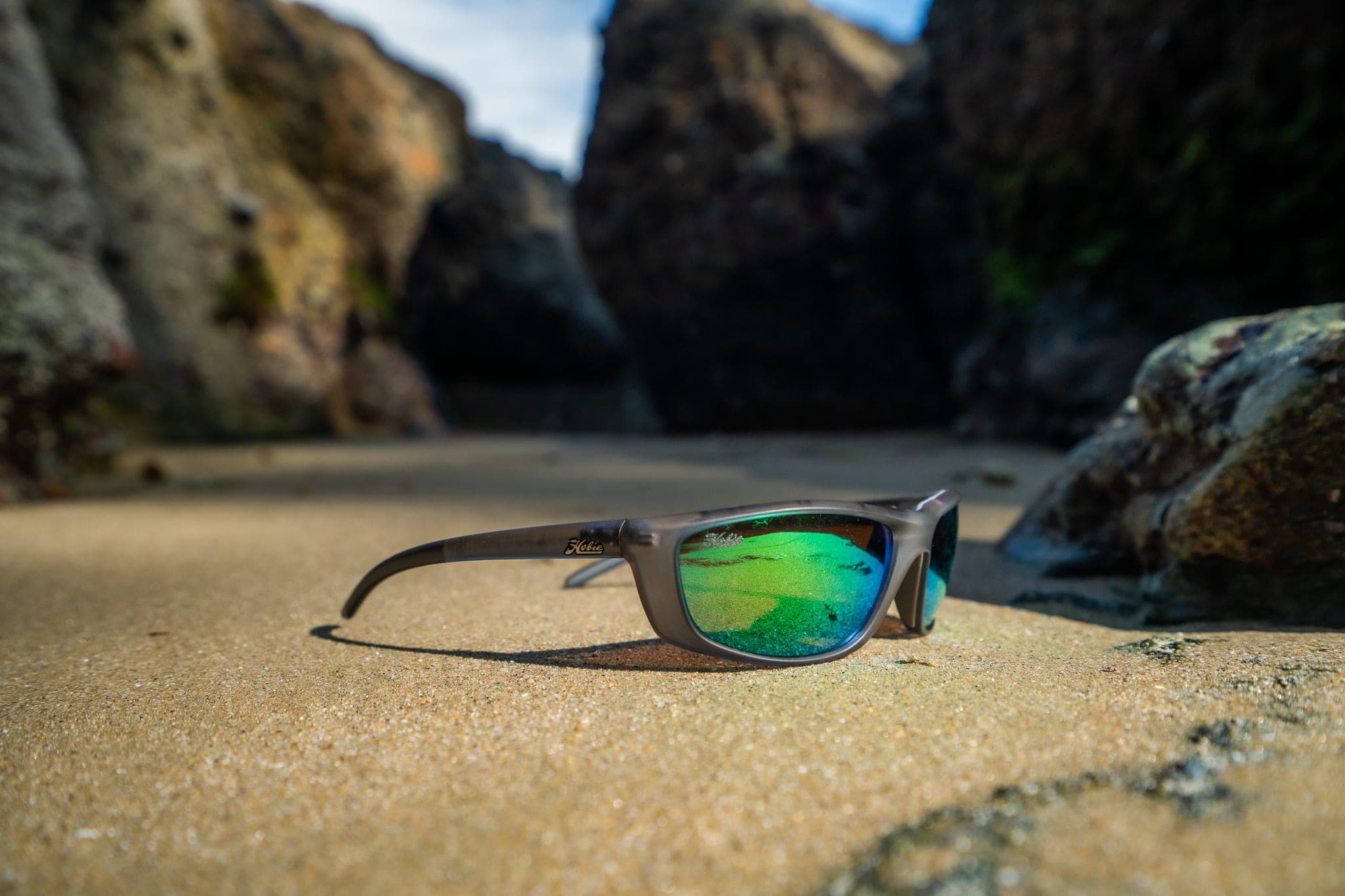 Green-mirrored sunglasses resting on wet sand between sunlit rocks at a beach cove