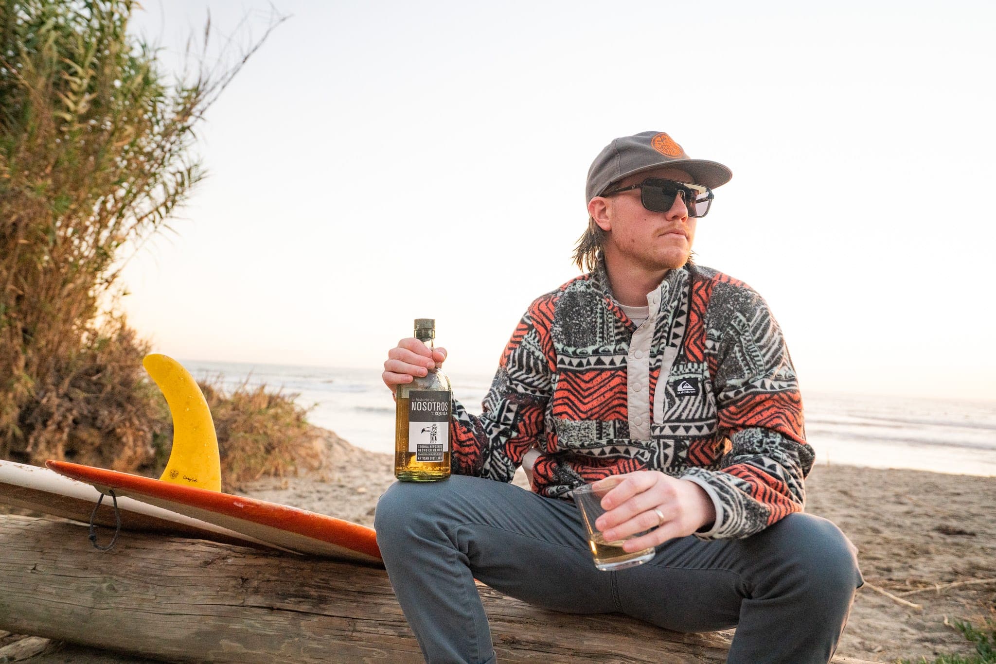 Man in a patterned fleece and cap sitting on driftwood with a bottle of tequila beside a longboard at sunset