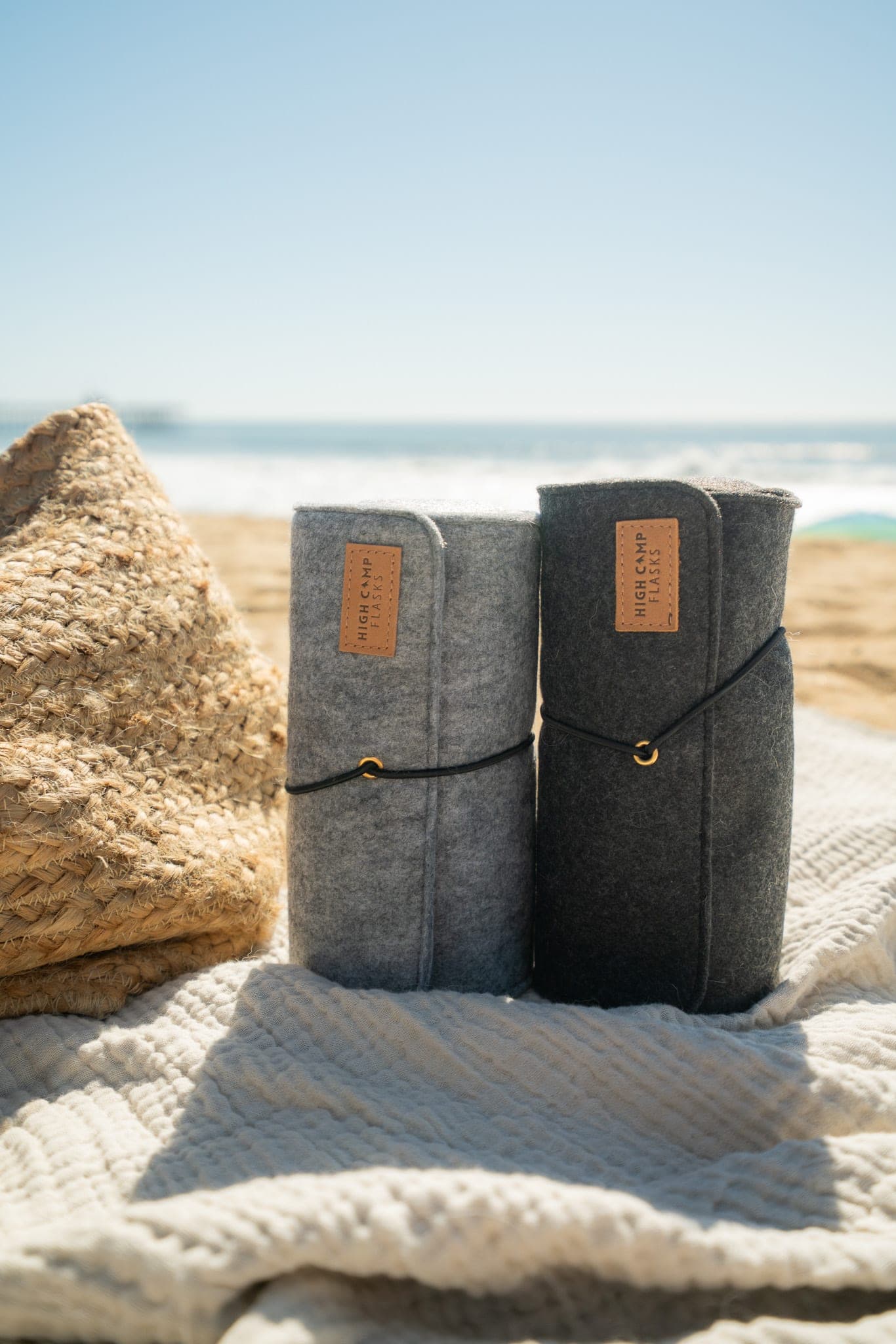 Two felt-wrapped flasks standing on a woven blanket with ocean waves in the background
