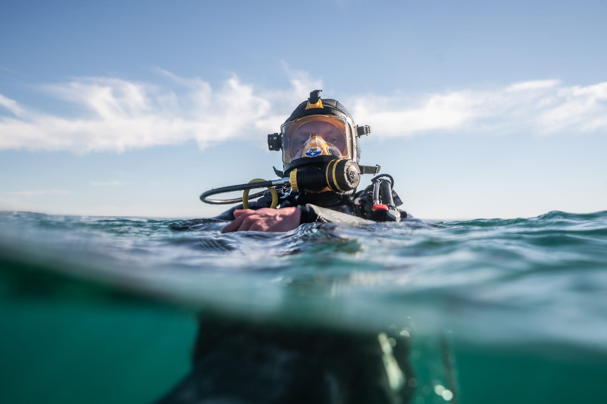Diver in a full-face mask surfacing in dark green ocean water under a wispy blue sky