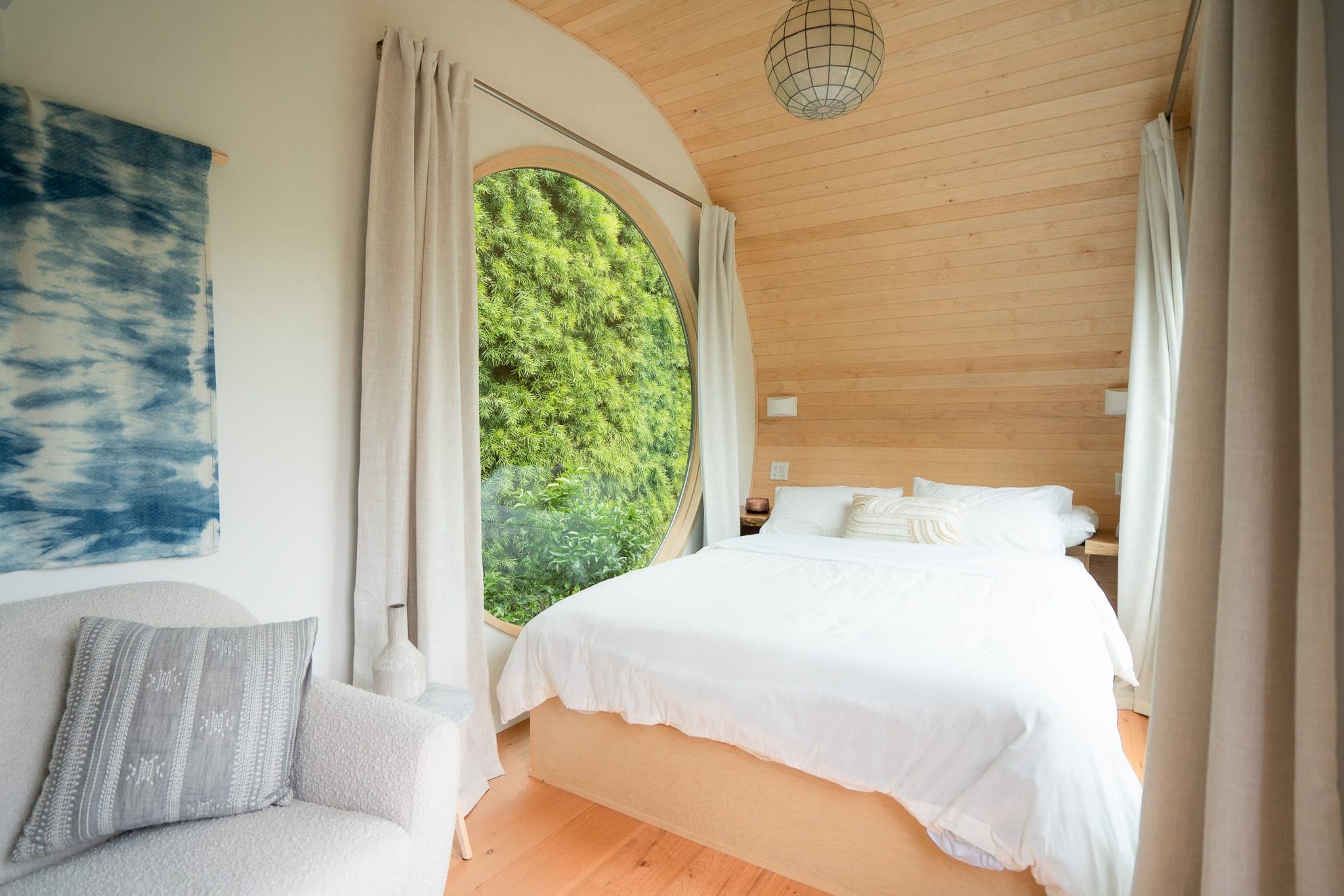 Cozy tiny-home bedroom with a large round window framing lush greenery beside a white bed