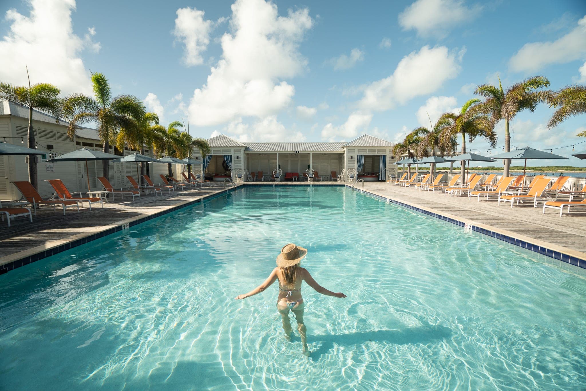 Woman in a straw hat wading into a palm-lined resort pool ringed with orange loungers