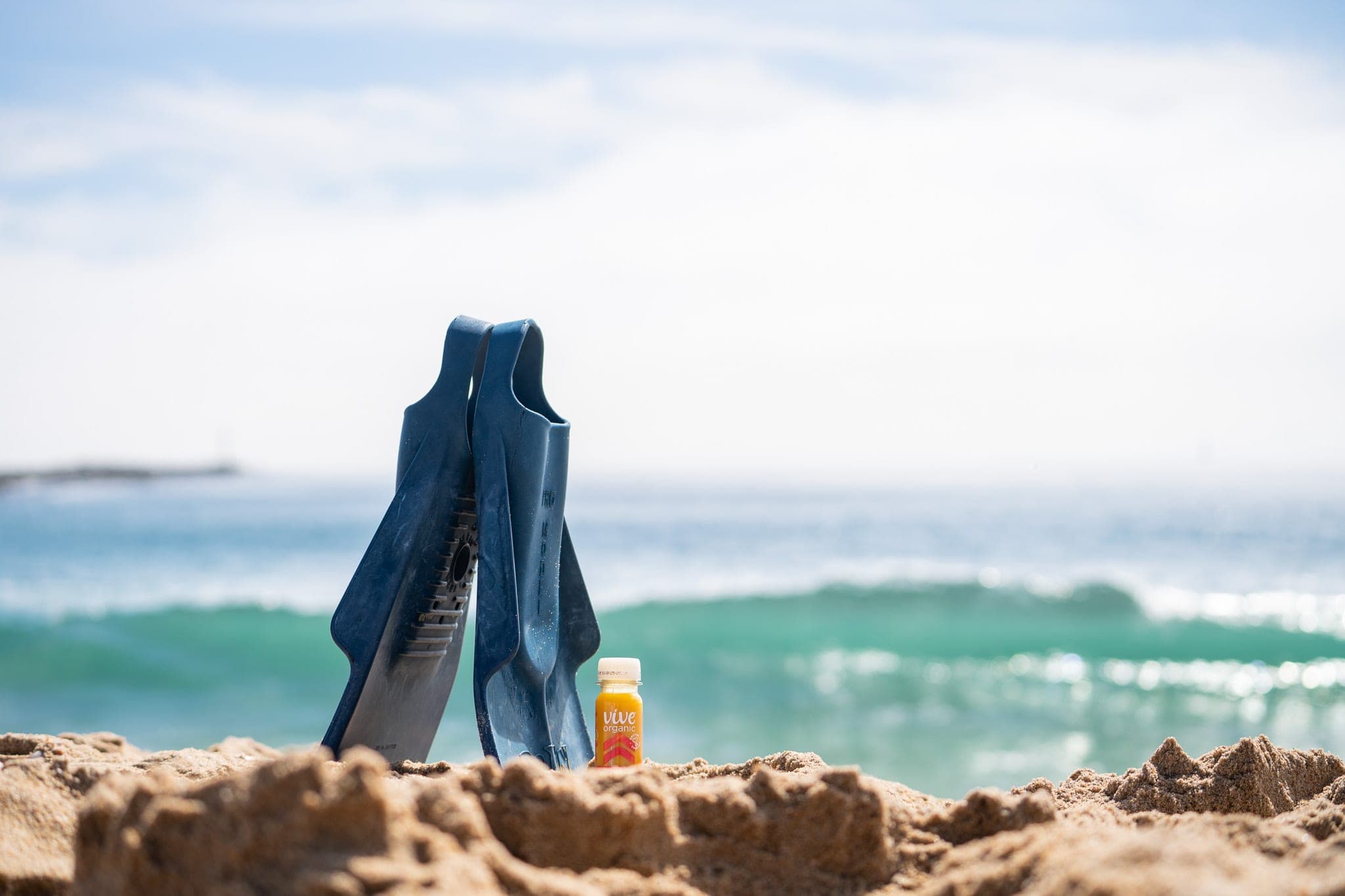 Pair of blue swim fins standing upright in the sand next to a juice shot with a wave breaking behind