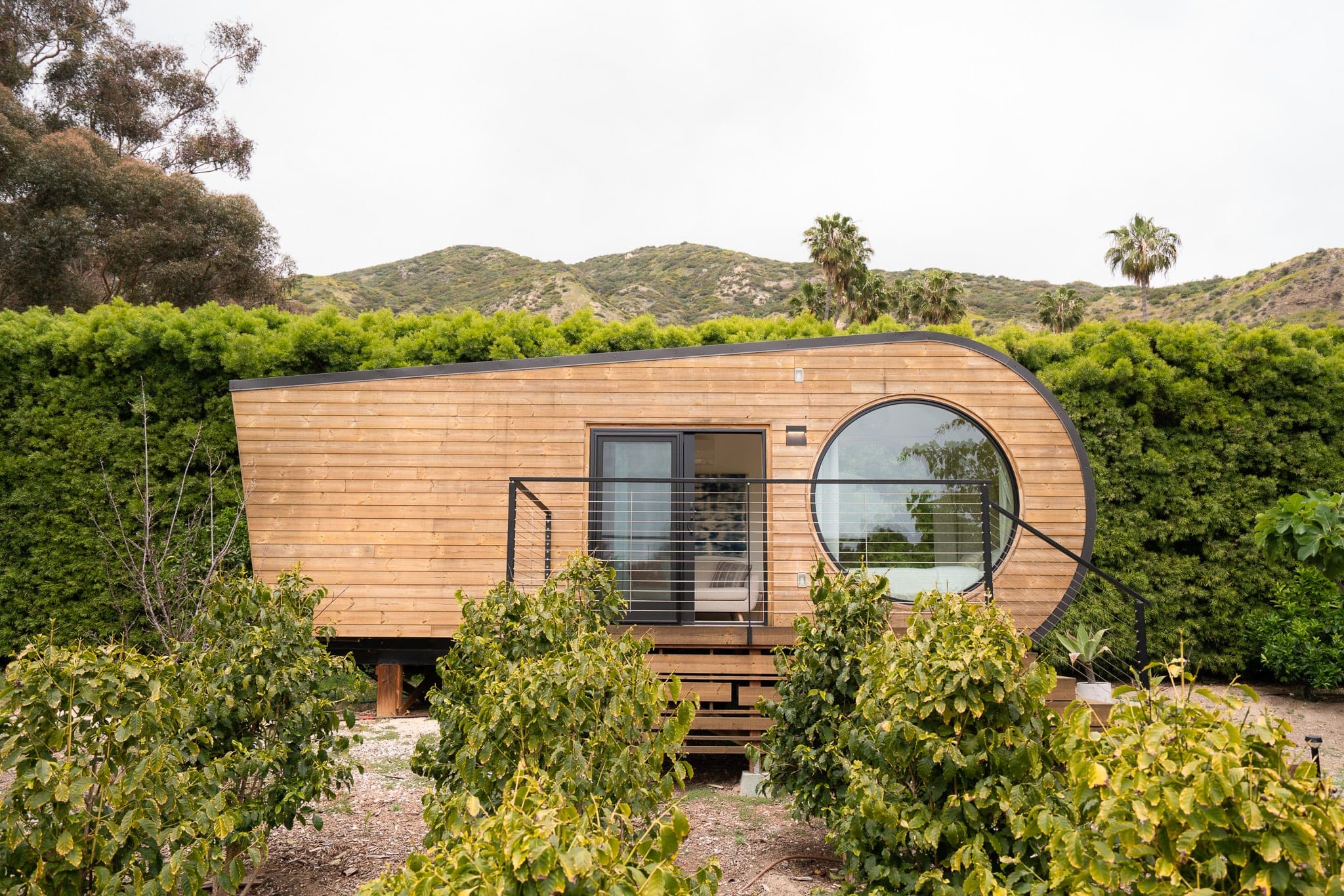 Curved wooden tiny house with a round porthole window set against a hedge and palm-dotted hills