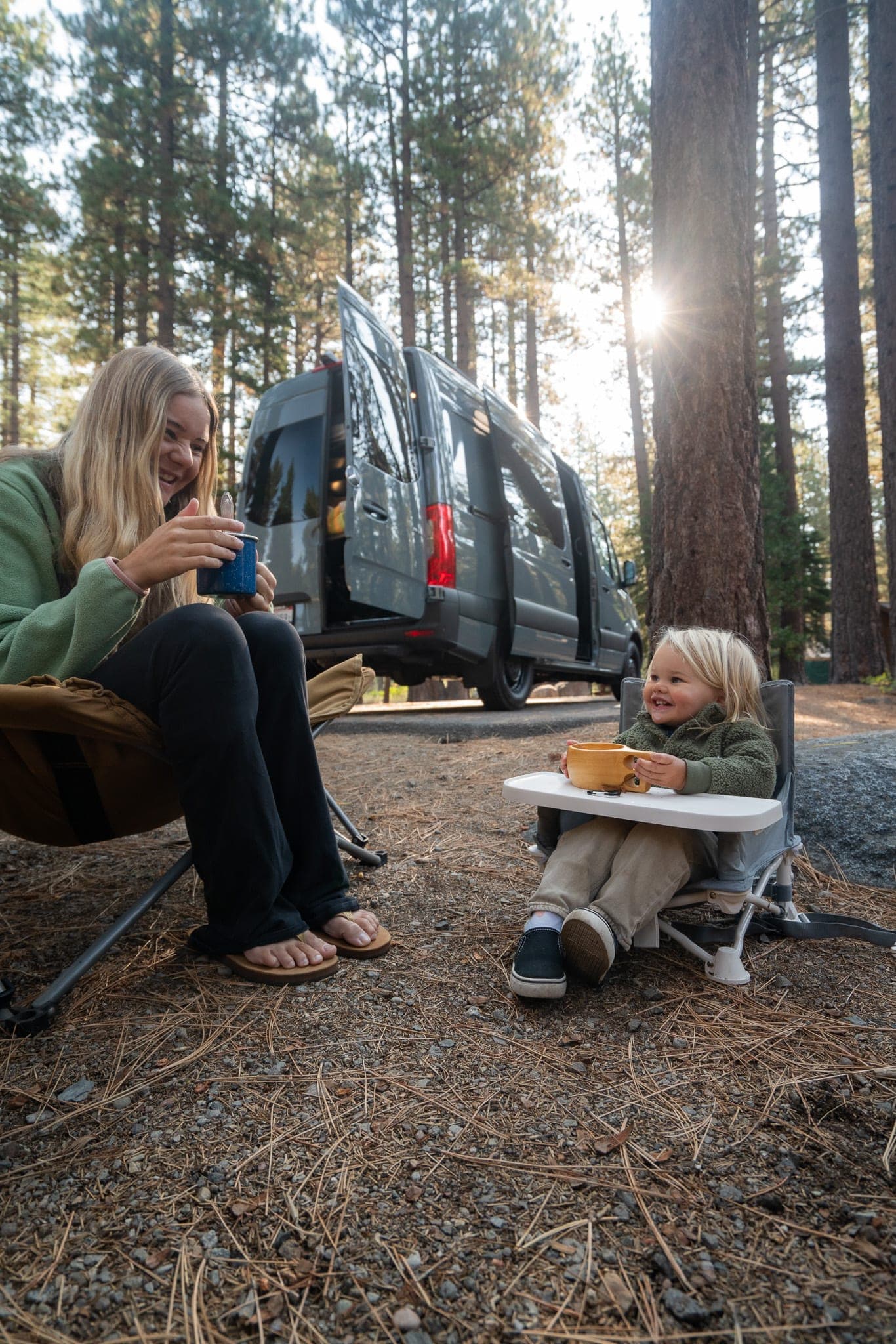 Mother and toddler sharing mugs in camp chairs beside a camper van in a sunlit pine forest